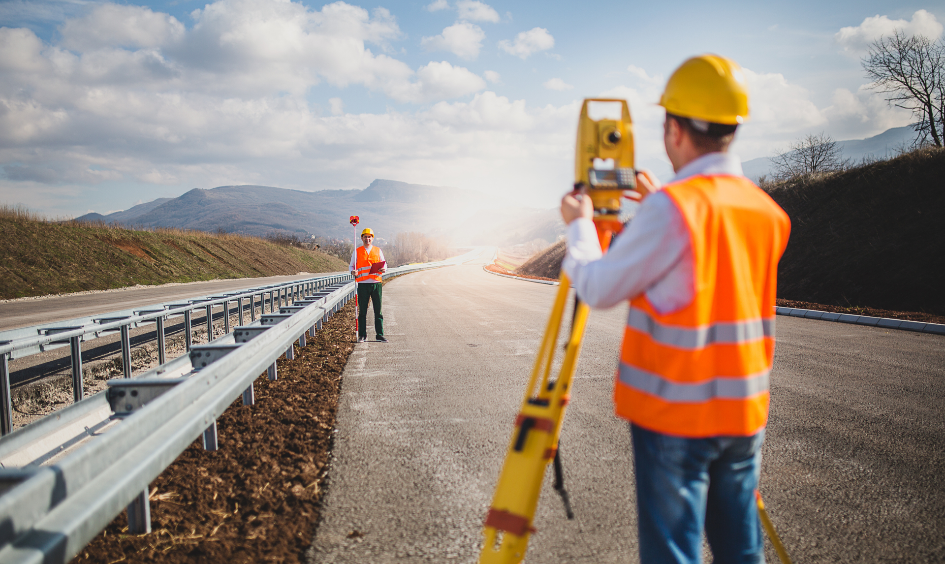 A worker in a high-visibility vest uses a surveying instrument on a road, with another worker holding a target nearby.