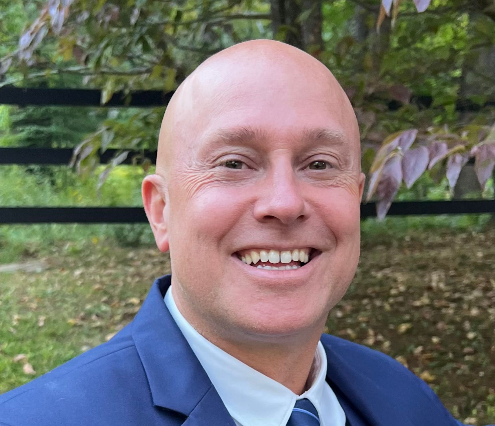 Bald man in blue suit smiles outdoors, against blurred foliage and fence.