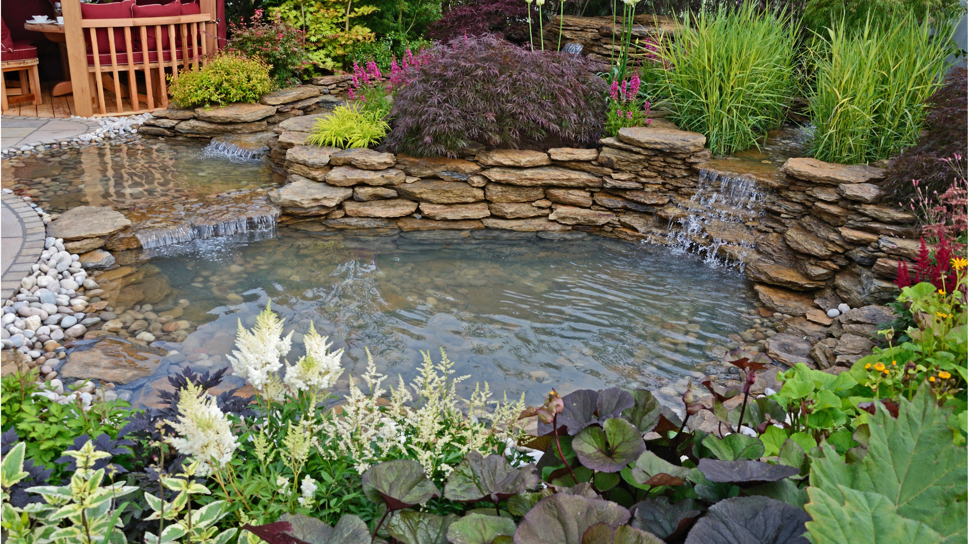 A pond with a waterfall in the middle of a garden surrounded by flowers.