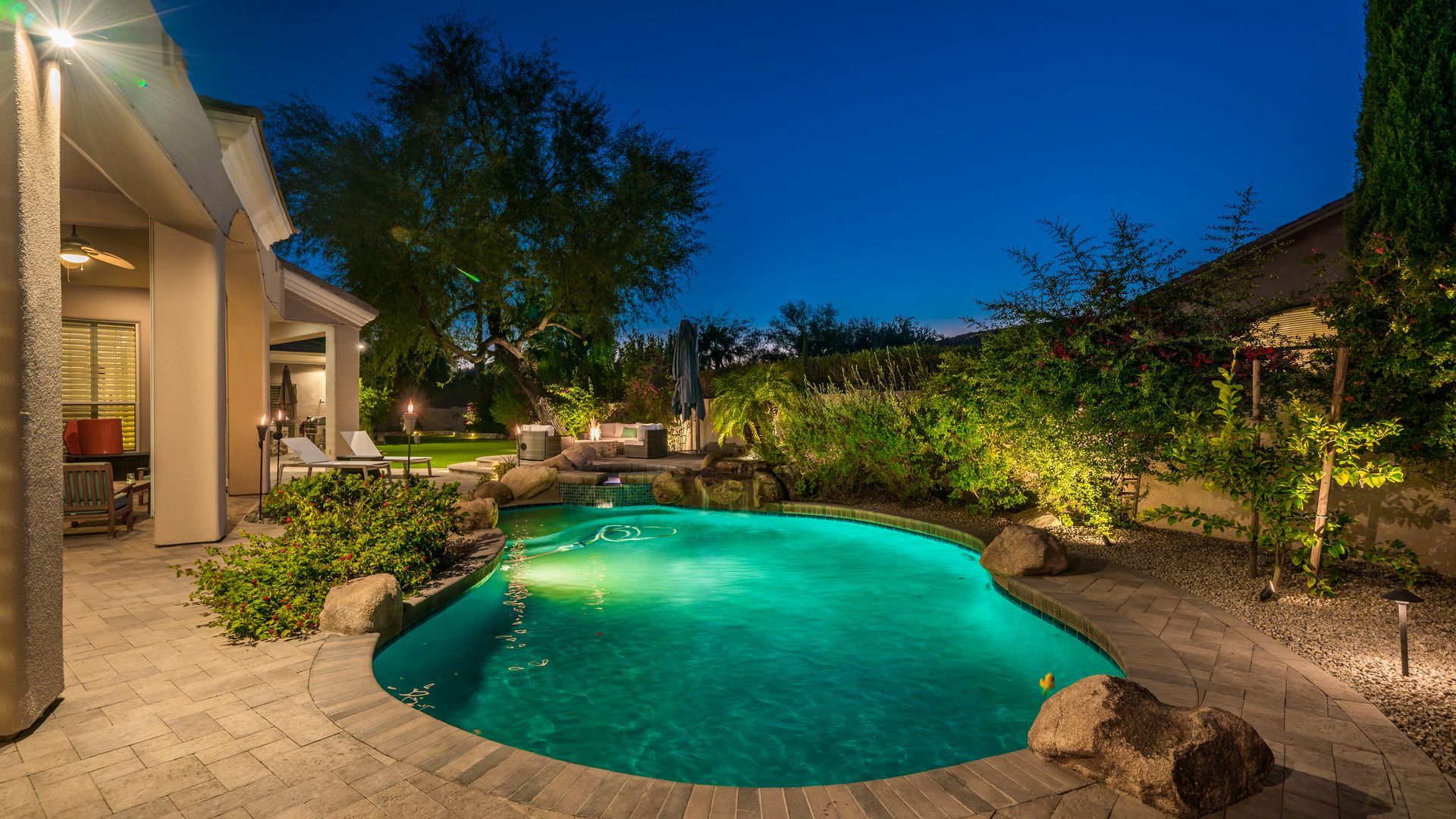 A large swimming pool is lit up at night in the backyard of a house.