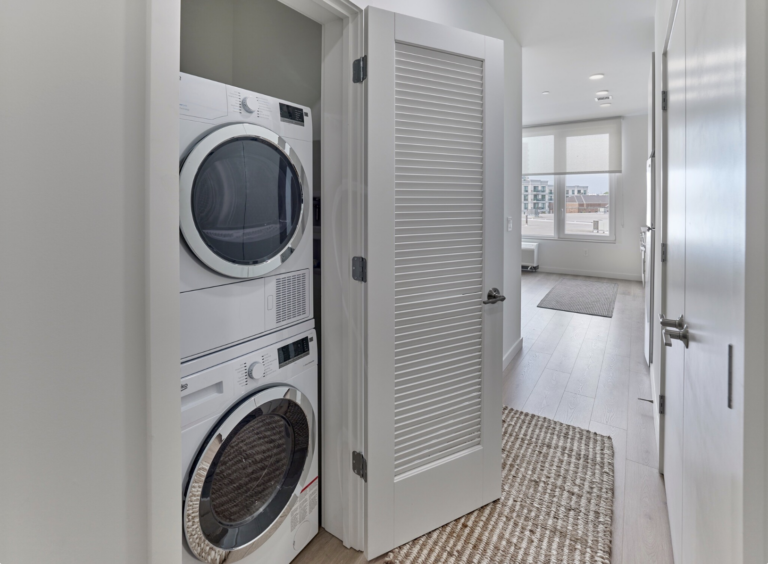 Stacked white washer and dryer in a narrow hallway. Ajar louvered door, neutral rug, and light wood floors.