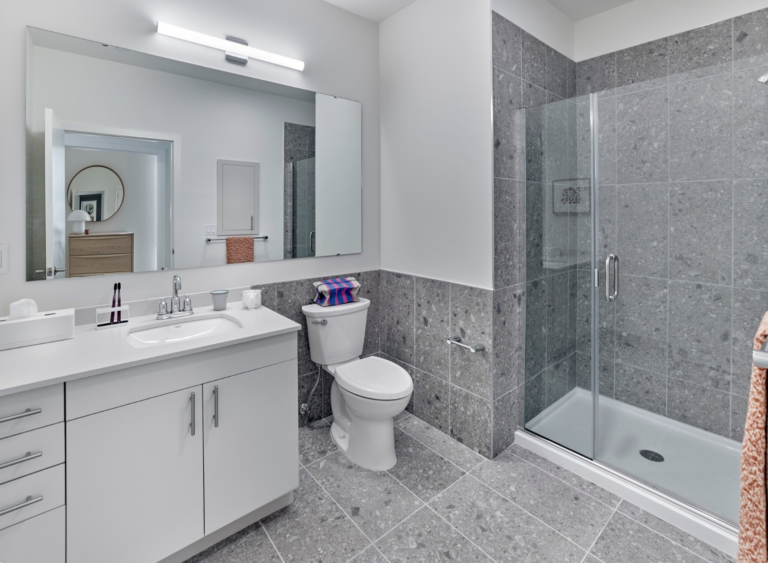 Modern bathroom with white vanity, gray tiled walls/floor, glass shower, and large mirror.
