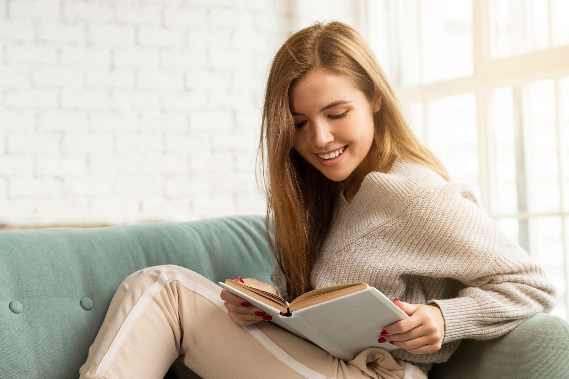 Woman sitting on a teal couch, smiling while reading a book. Natural light from a window.