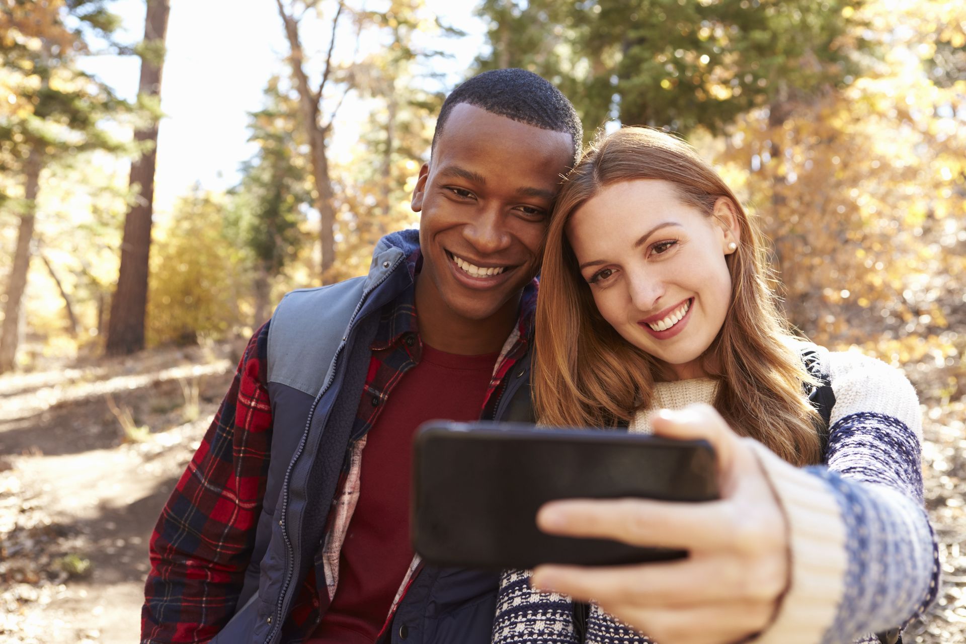 A smiling couple takes a selfie outdoors among trees. Man wears a vest and plaid shirt. Woman in a patterned sweater.