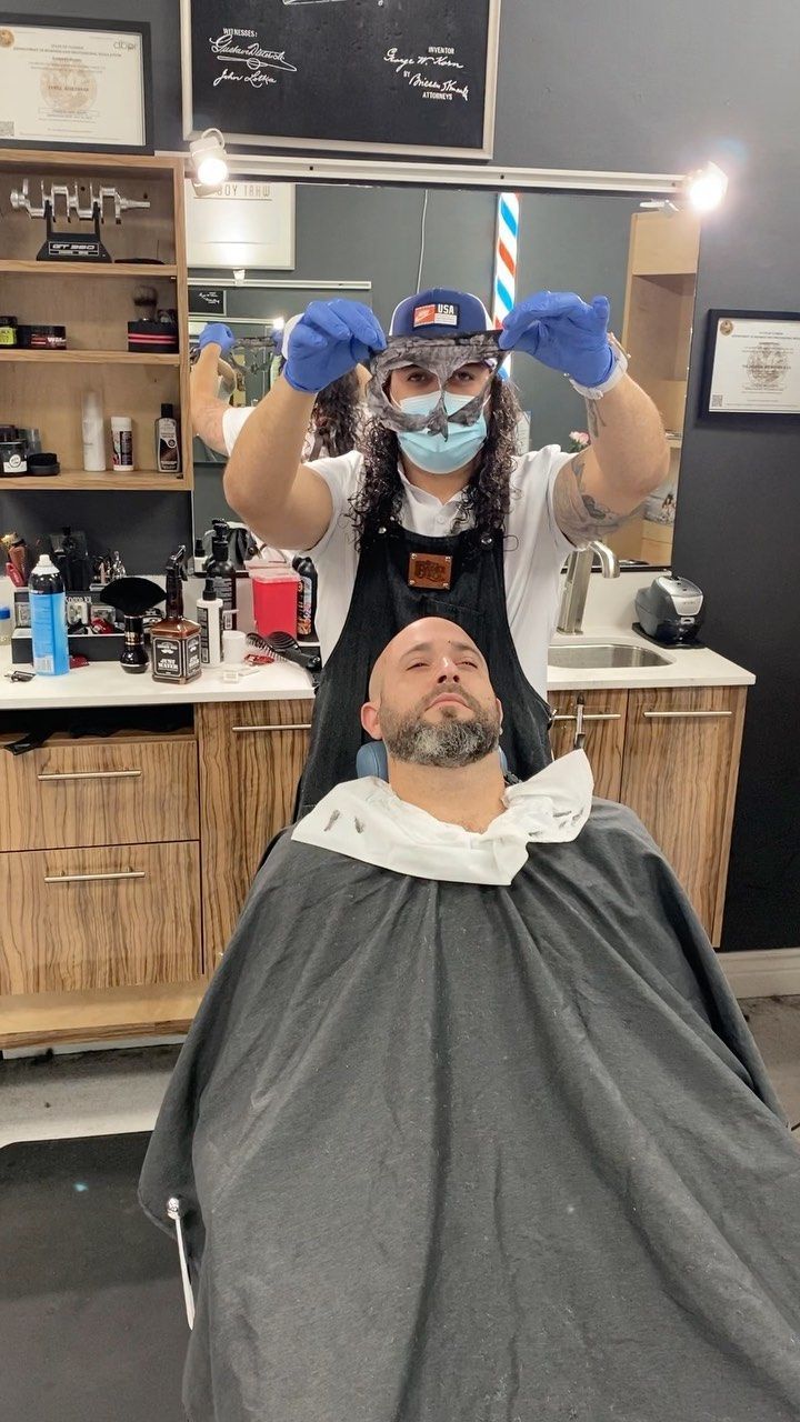 Barber shop scene: masked stylist holds clippers above a client reclining in a barber cape.