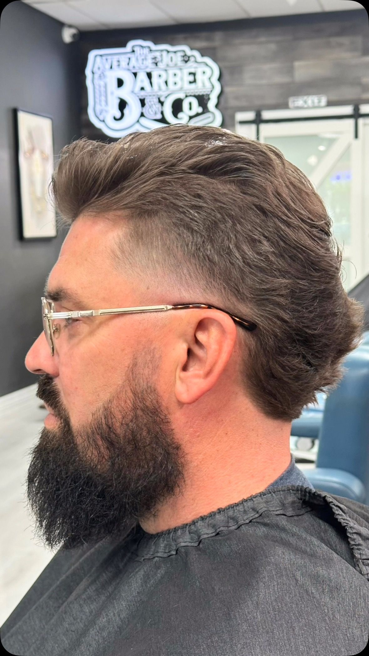 Profile view of a man with glasses and a full beard, seated in a barber shop with a styled haircut.