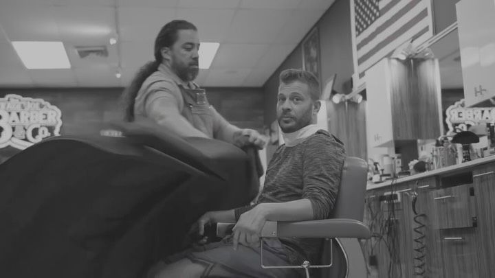 Two people in a barbershop, one seated in a barber chair, with mirrors and grooming tools nearby.