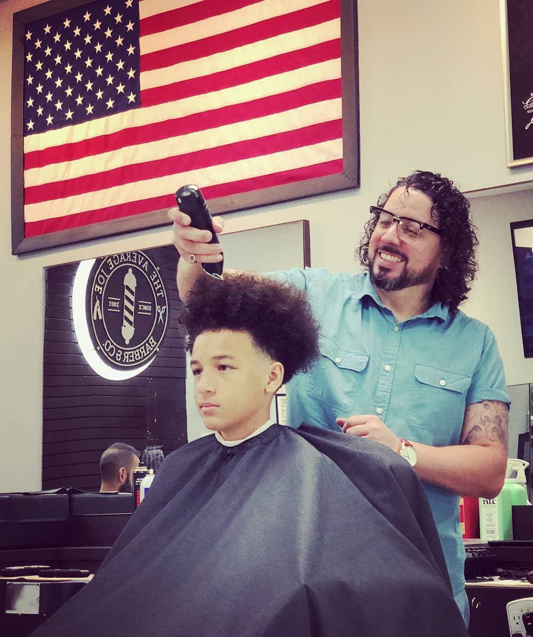 Barber cutting a client's hair in a shop, with an American flag on the wall.