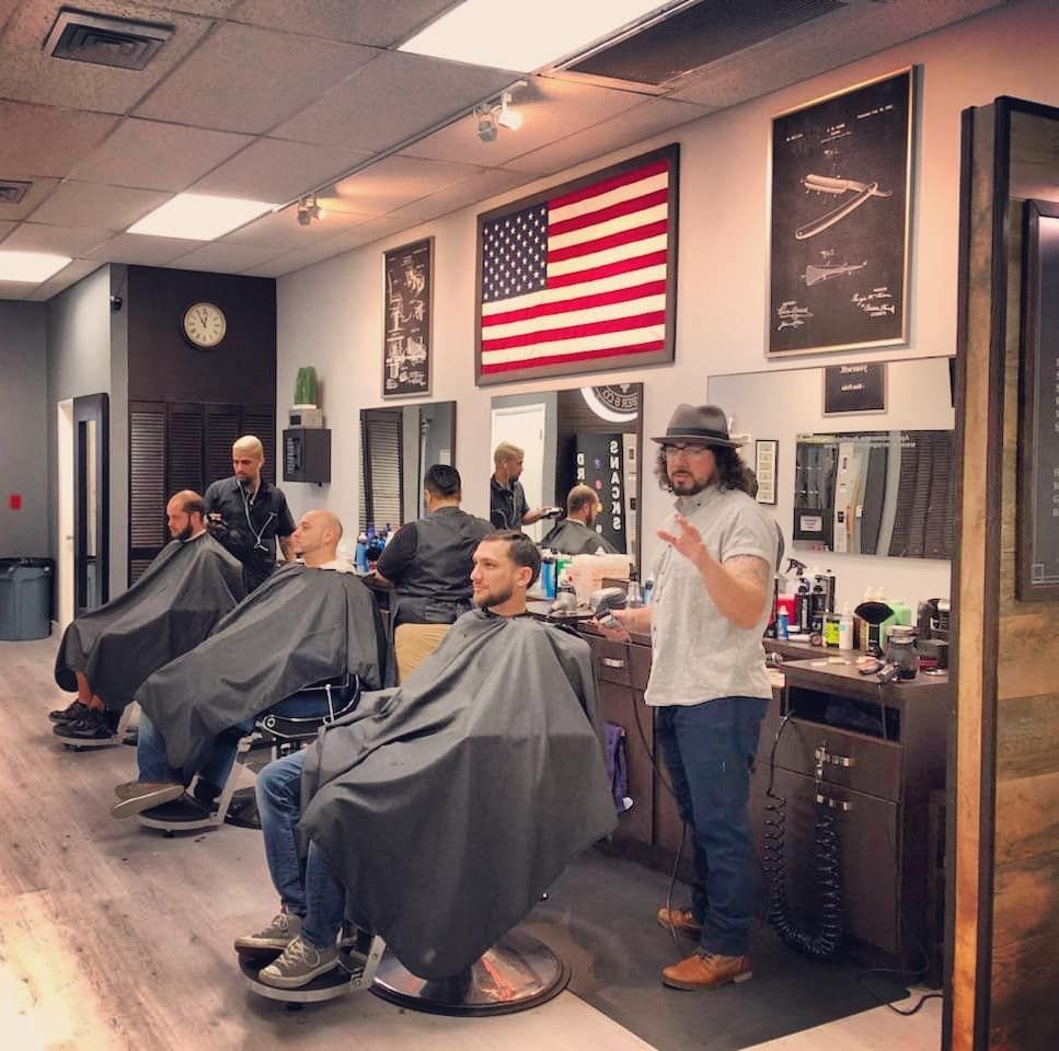 Barbershop interior with three clients in capes, a barber standing by the mirror, and a large American flag on the wall