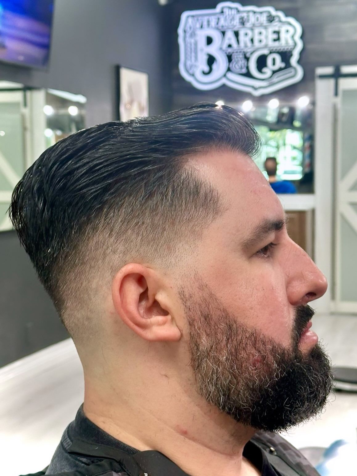 Side-profile haircut in a barbershop, with a neat fade and trimmed beard under a lit barber sign.