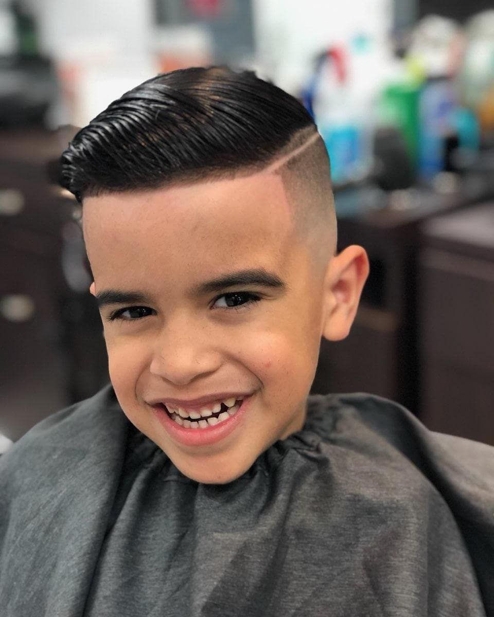 Smiling child with slicked-back hair and braces, seated in a barber chair wearing a gray cape.
