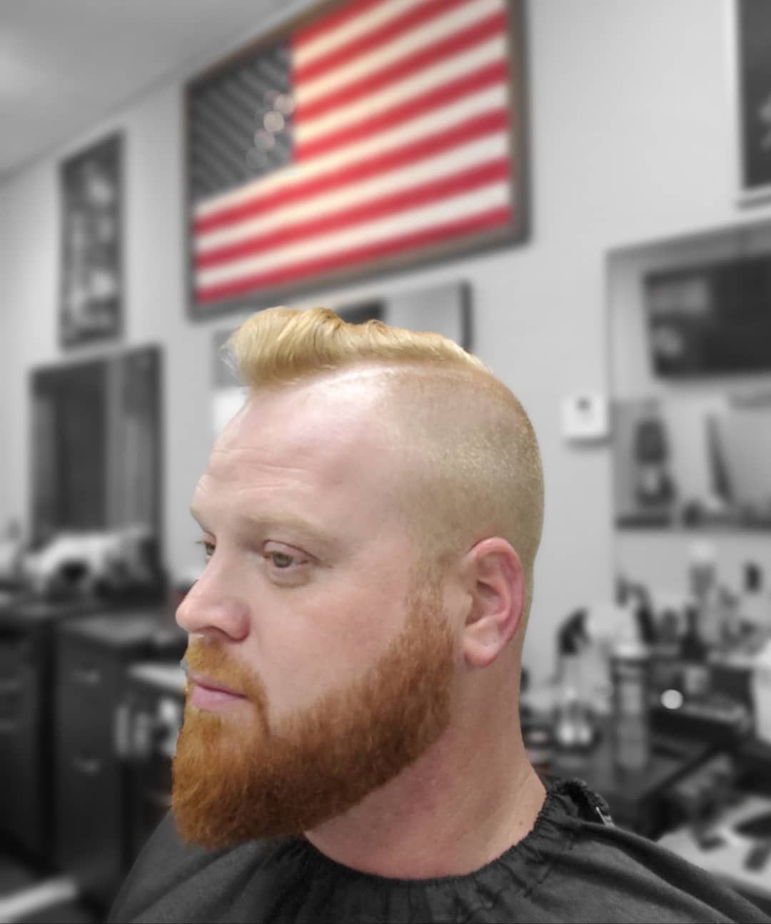 Profile view of a red-bearded man in a barbershop, with an American flag on the wall behind him