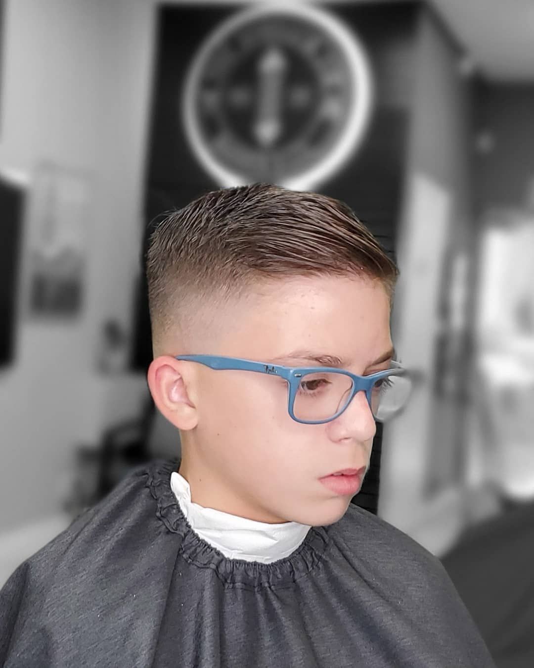 Child with blue glasses and short haircut in a barbershop, wearing a gray cape and white neck strip.