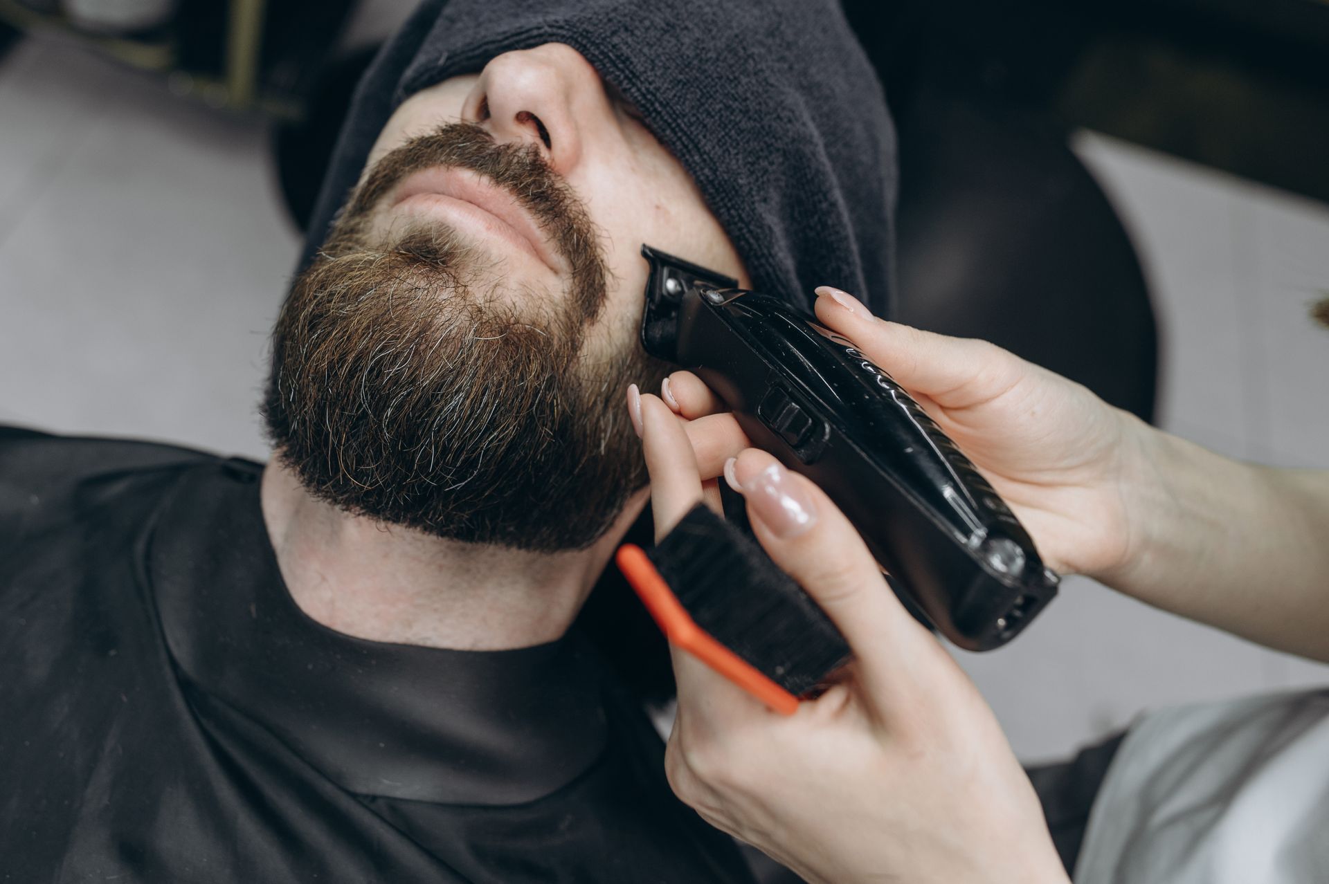 Bearded man getting a beard trim with electric clippers while wearing a black beanie