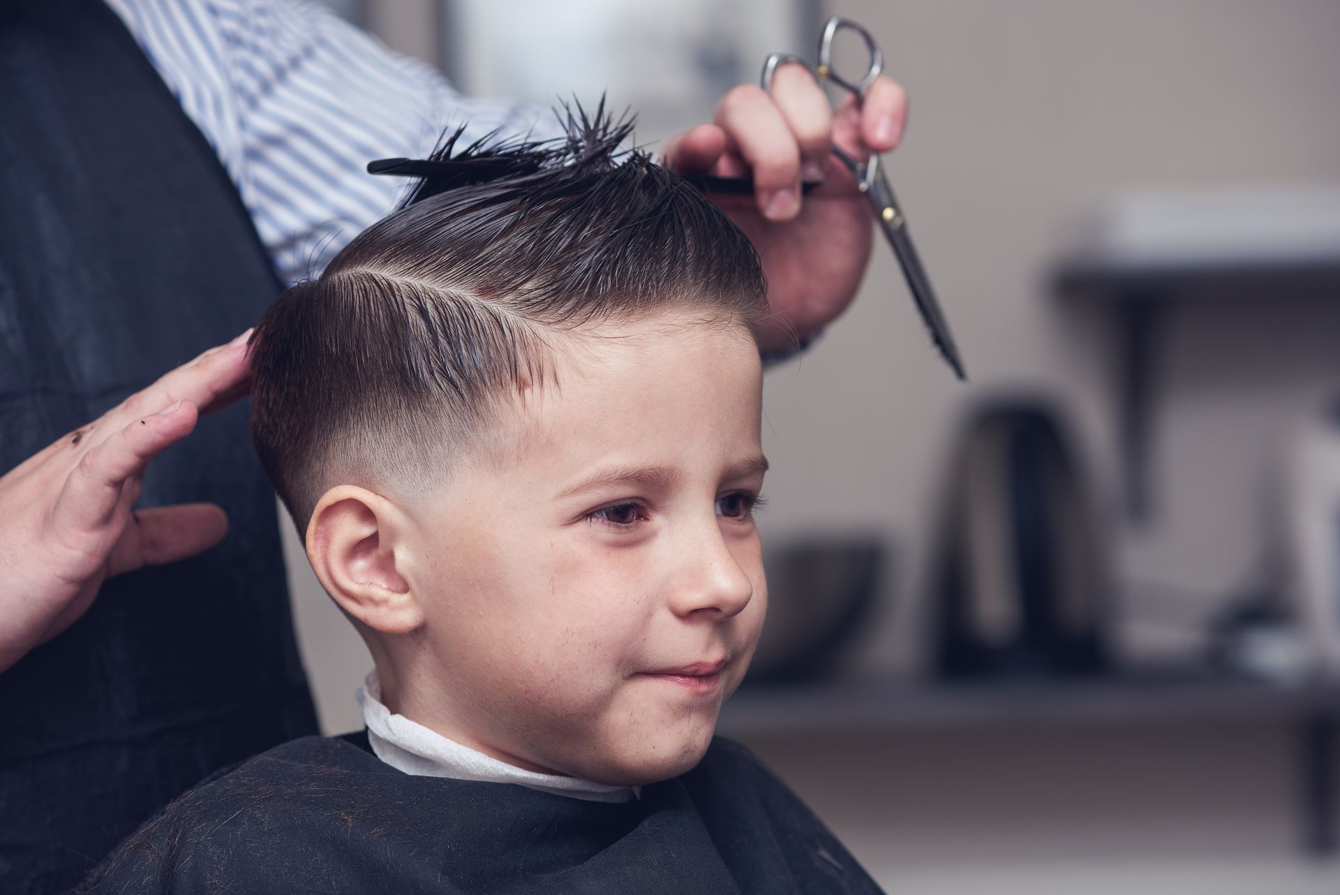 Haircut in progress with scissors as a child sits in a barbershop cape, smiling slightly.