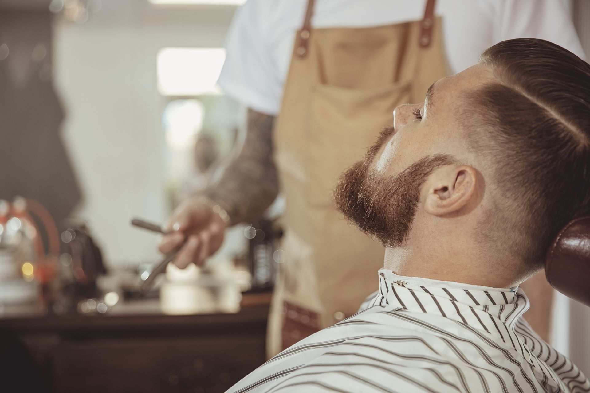 Man reclining in a barber chair while a barber trims his beard in a bright barbershop