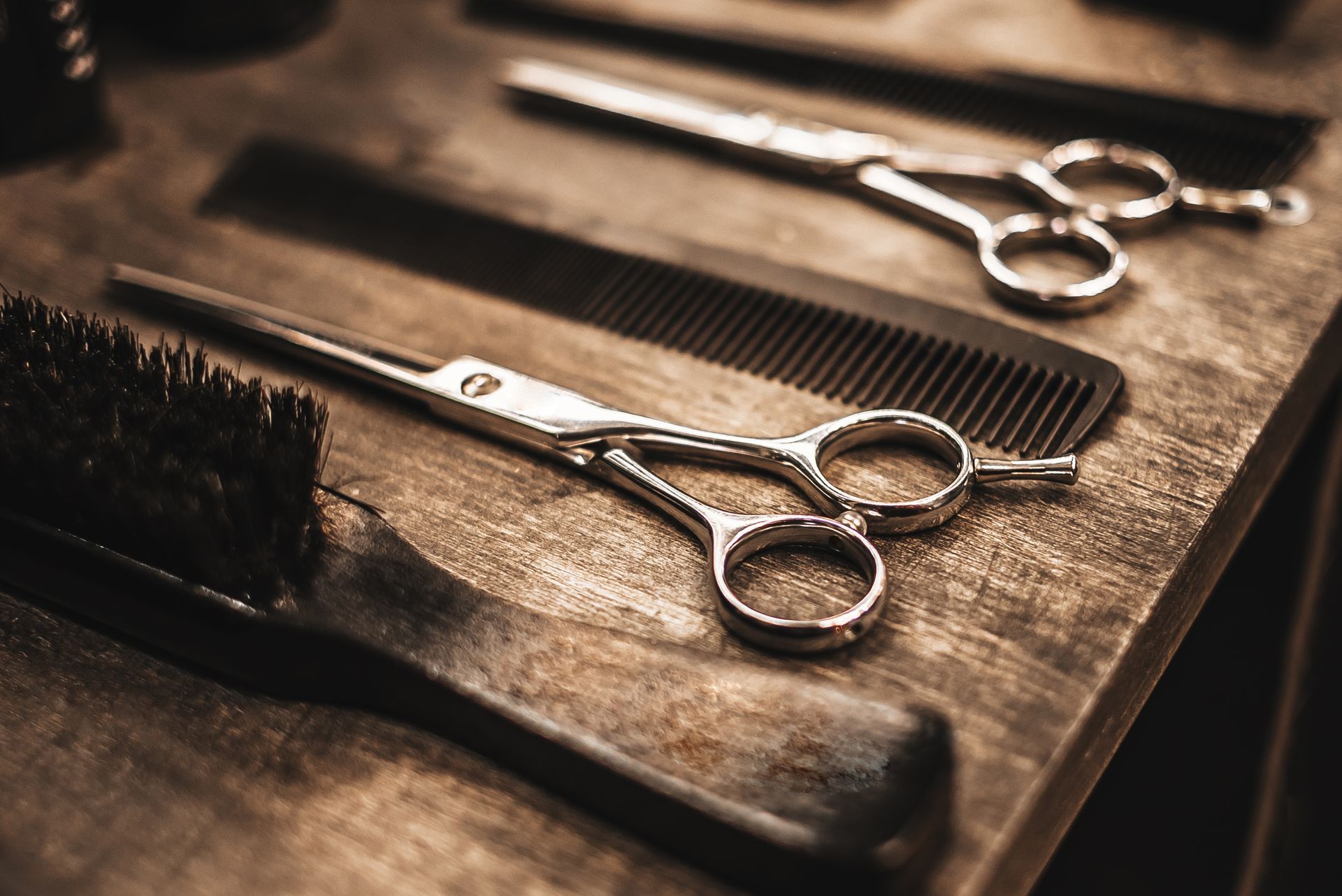 Two silver barber scissors and combs on a wooden tabletop, lit warmly from above