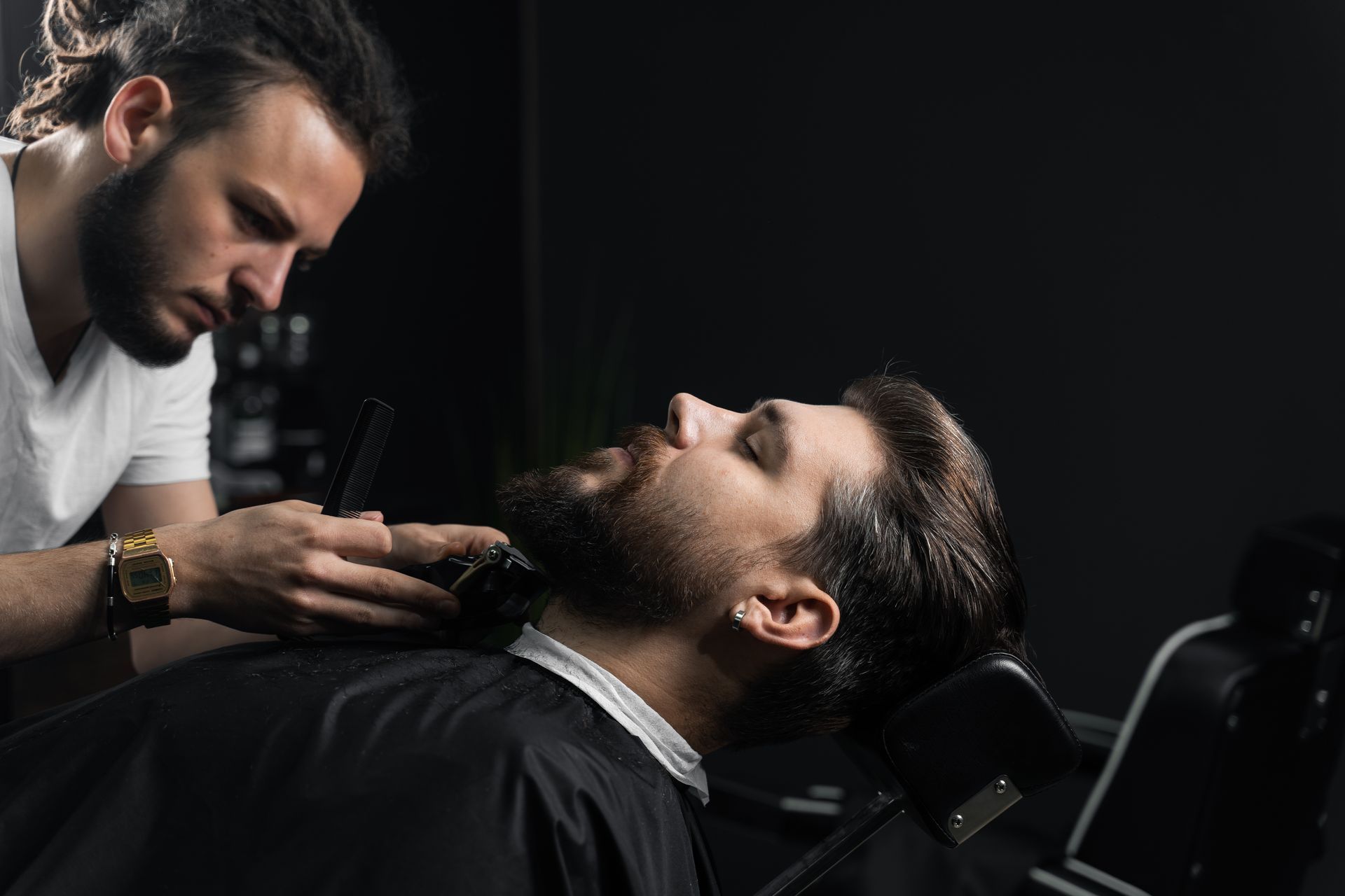 Barber trimming a man’s beard in a dark barbershop, the client reclined in the chair.