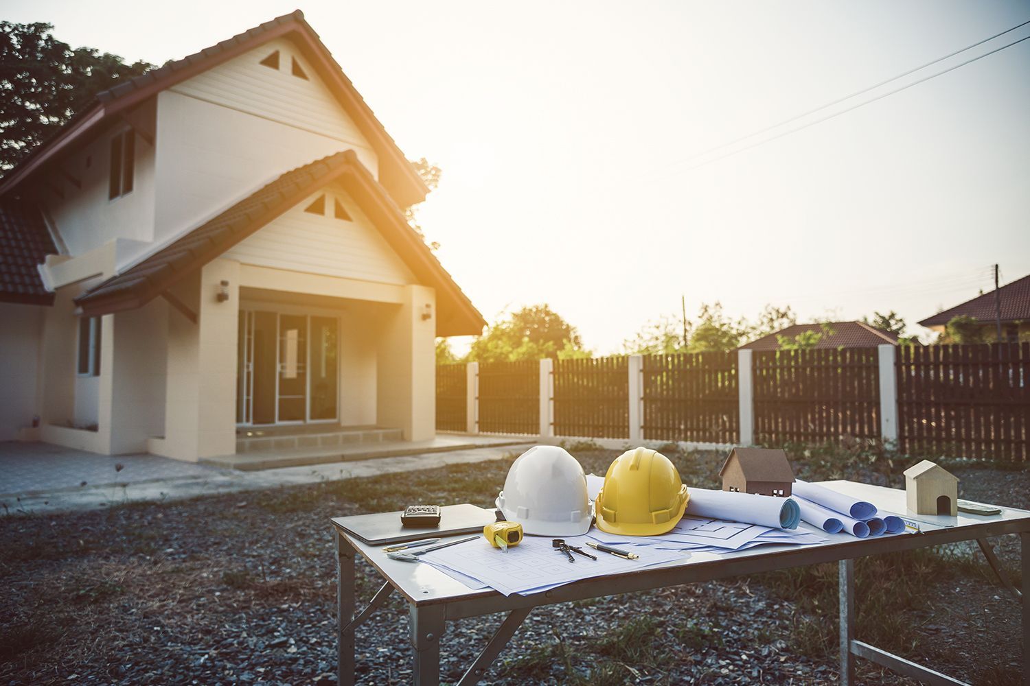 Construction table with blueprints and hard hats in front of a house under renovation.