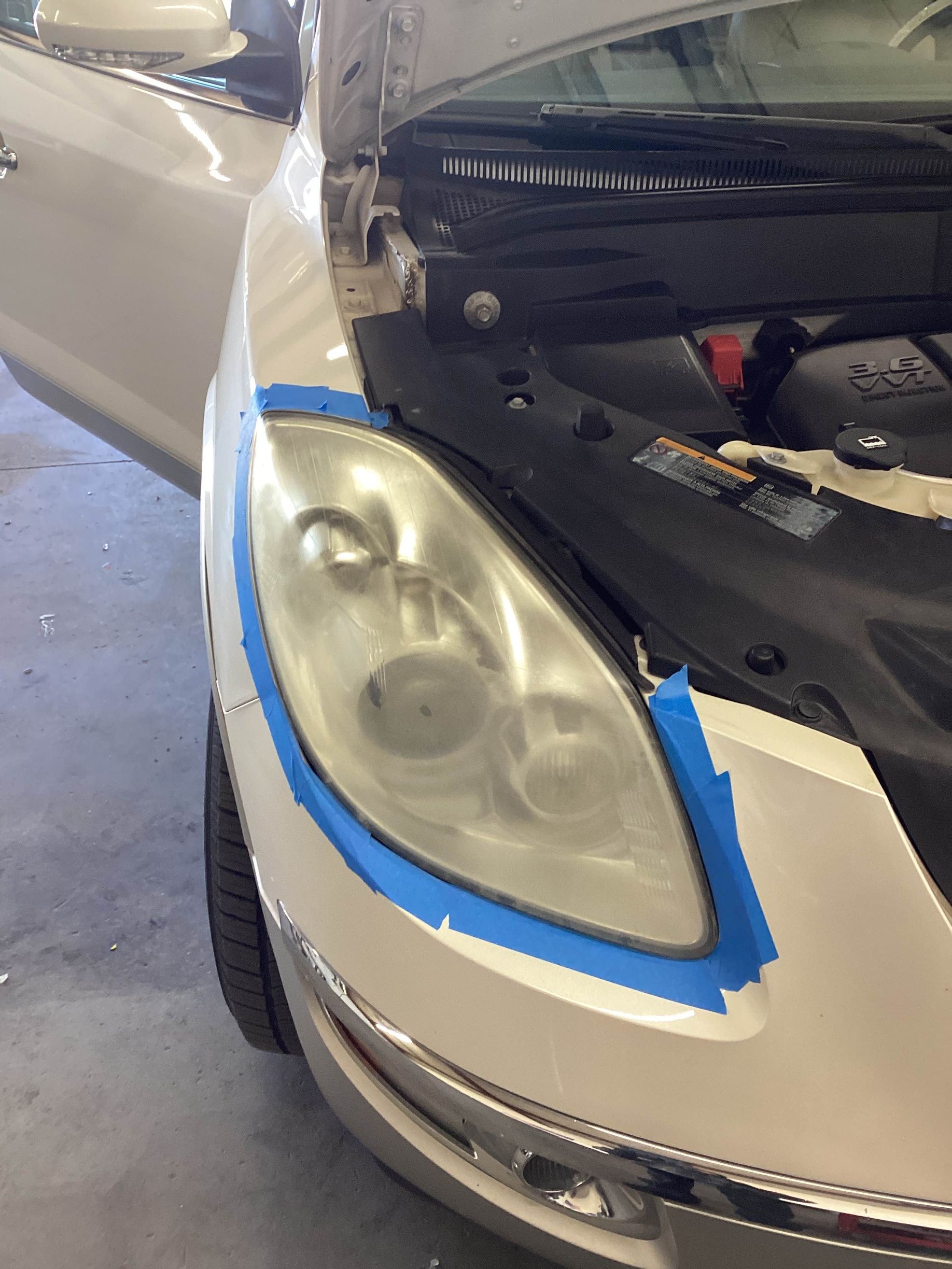 Car headlight being restored, masked with blue tape. The hood and door are open.