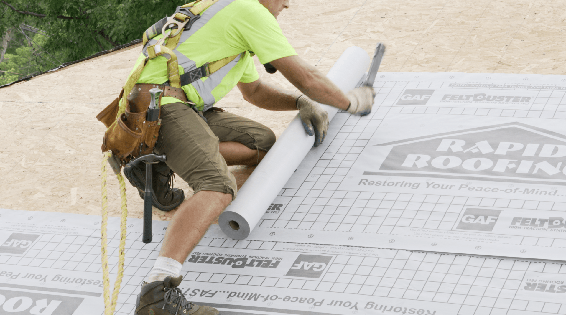 A man is kneeling on the ground holding a roll of roofing paper.