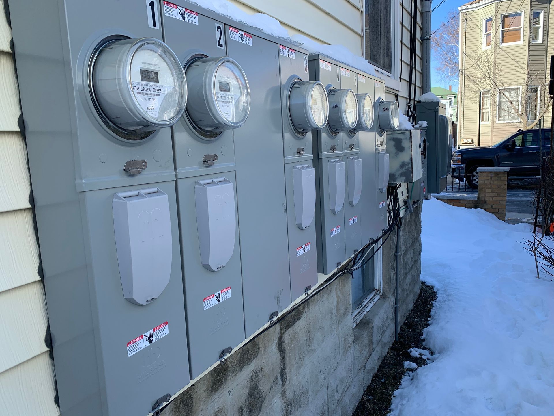 Exterior view of gray electrical meters on a building, with snow on the ground and a city street in the background.
