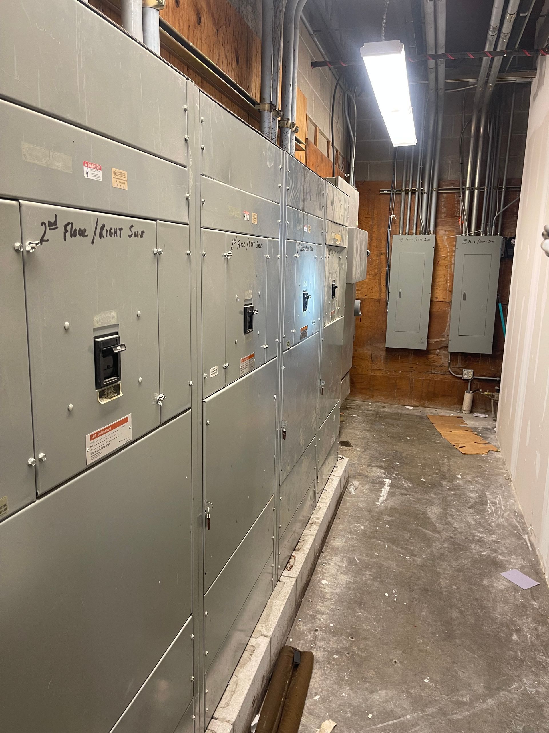 Electrical panels line a narrow corridor; grey metal cabinets against a brick and concrete wall.