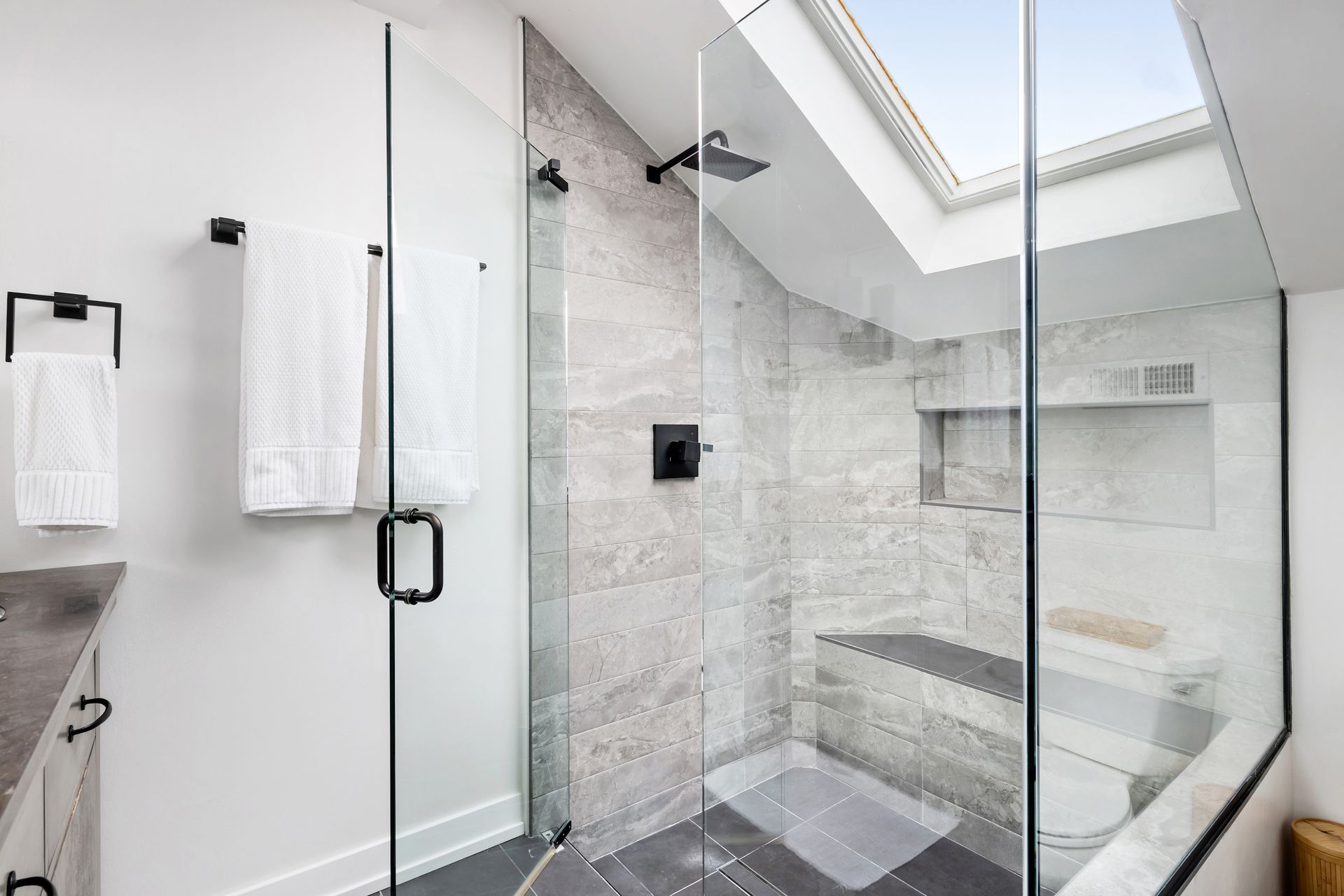 Modern bathroom with glass shower enclosure, gray tile, and skylight overhead