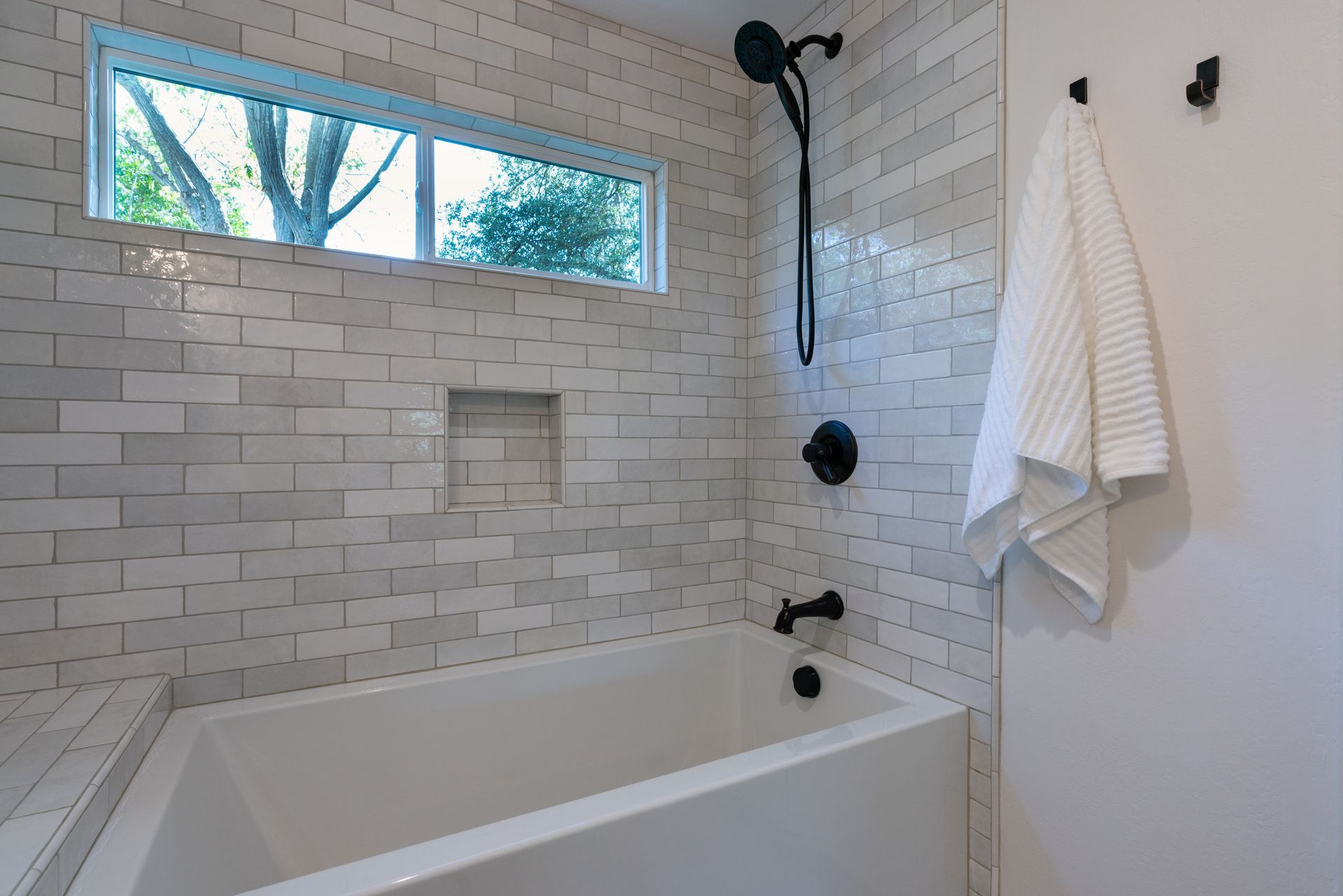 Modern white-tiled bathroom with a bathtub, shower fixtures, window, and hanging towel