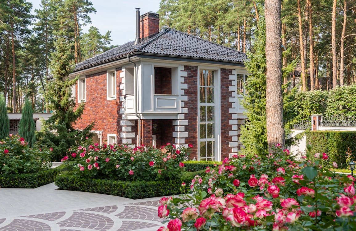 Brick house with white trim, surrounded by rose bushes, on a paved driveway in a wooded area.