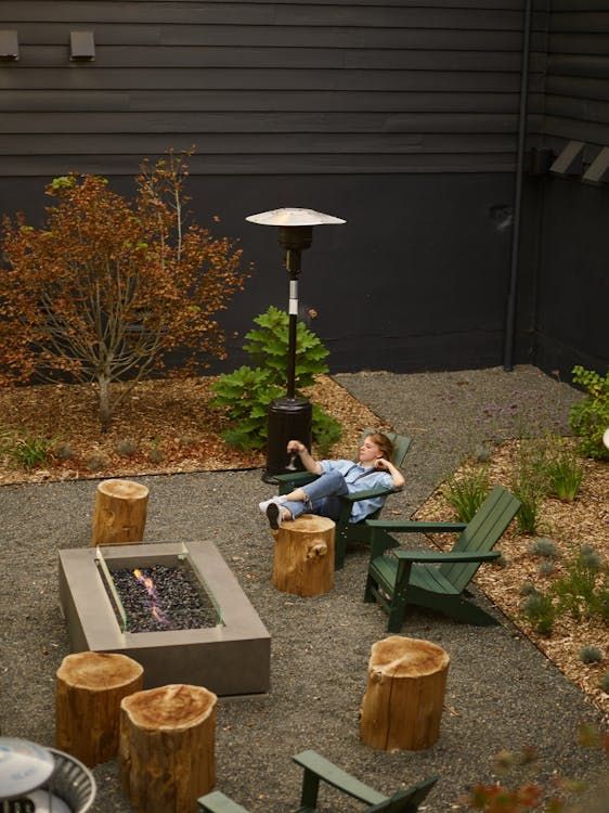 Person relaxing in a backyard with a fire pit, stumps, and a patio heater.