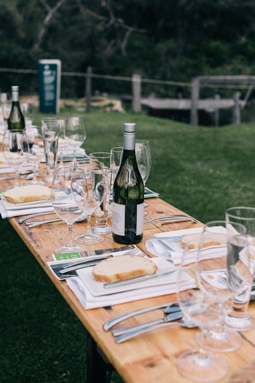 Outdoor table set for a meal with wine bottles, glasses, plates.