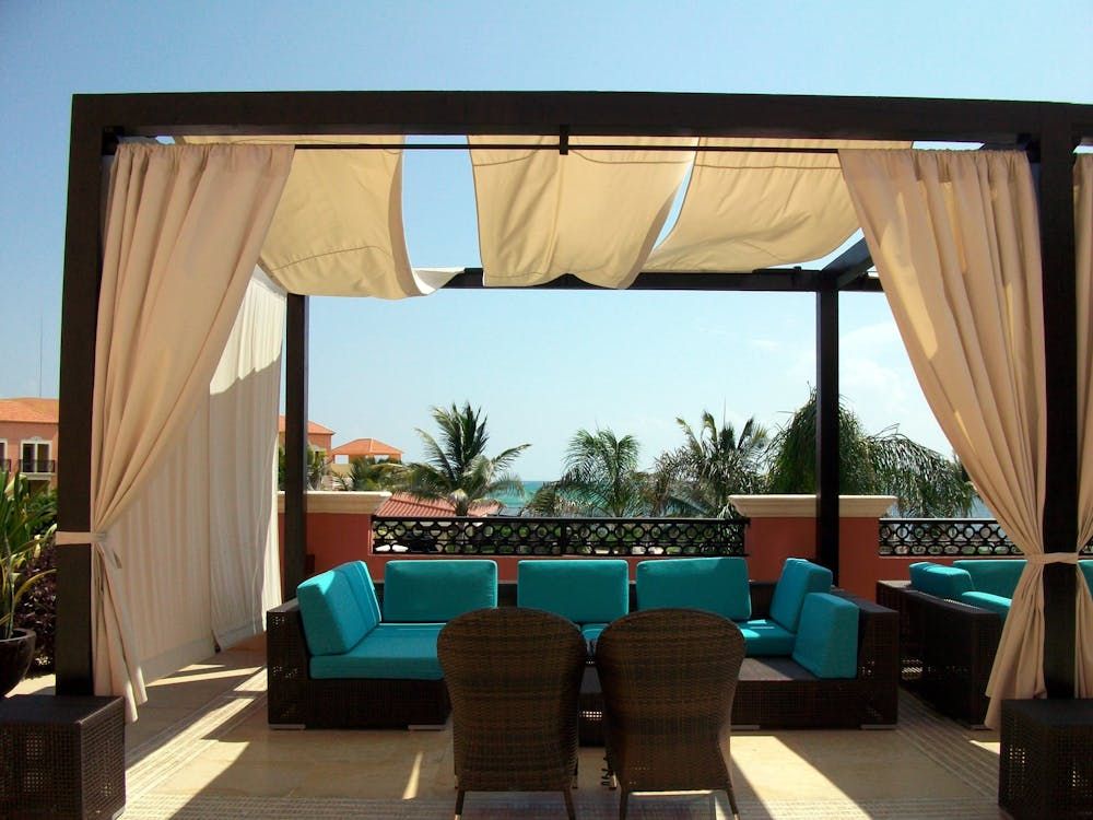 Outdoor lounge area with turquoise cushions, tan curtains, and a view of the ocean.