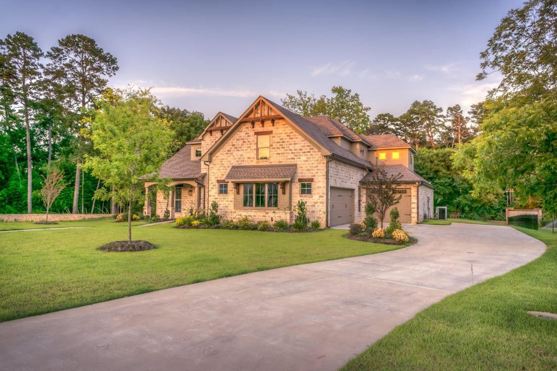 Stone-clad house with a long driveway on a grassy lot, surrounded by trees under a blue sky. Stone-clad house with a long driveway on a grassy lot, surrounded by trees under a blue sky.
