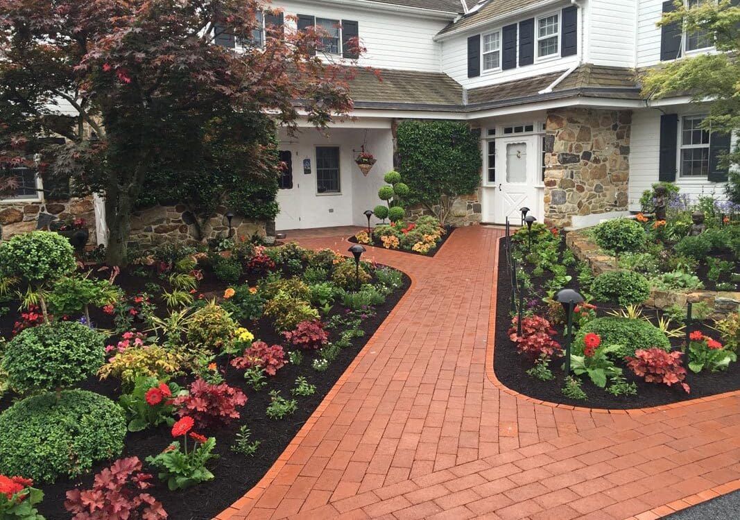 Brick pathway leads to a white house with stone accents, flanked by vibrant flowerbeds with red and green plants. Brick pathway leads to a white house with stone accents, flanked by vibrant flowerbeds with red and green plants.