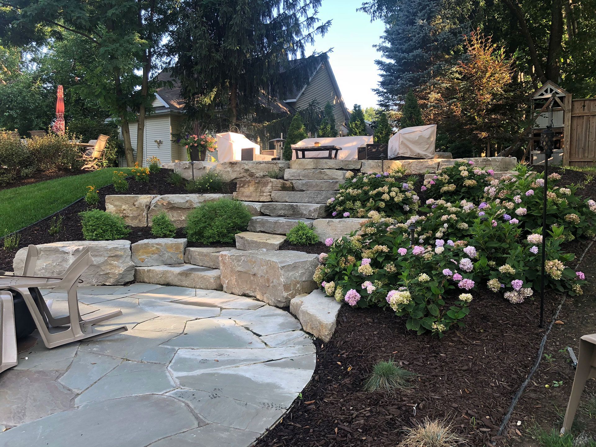 Stone patio and stairs lead to a seating area in a landscaped backyard with flowering bushes.
