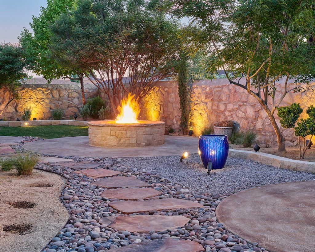 Fire pit in a stone-walled backyard with a stepping stone pathway, gravel, and illuminated trees. Fire pit in a stone-walled backyard with a stepping stone pathway, gravel, and illuminated trees.