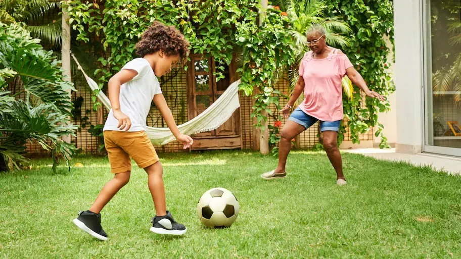 A child kicking a soccer ball while playing with an older person in a grassy backyard.