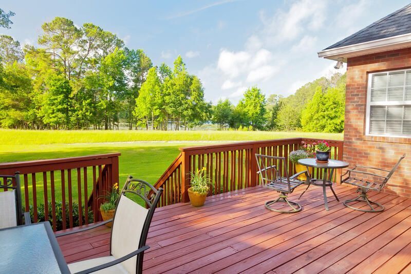 Wooden deck with outdoor furniture overlooking a grassy field and trees under a sunny sky. Wooden deck with outdoor furniture overlooking a grassy field and trees under a sunny sky.