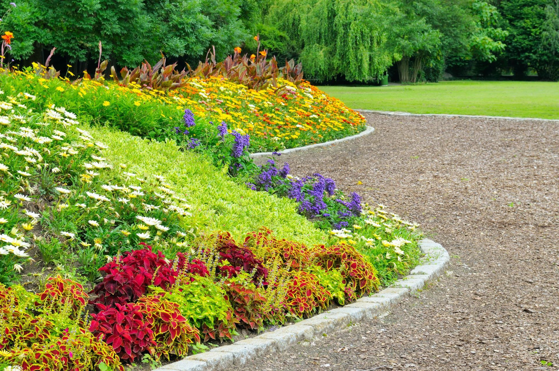 Colorful flower bed with curved stone border and gravel path in a sunny park.