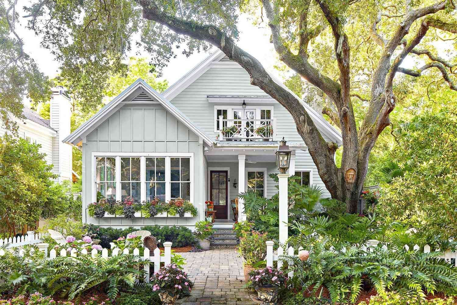 Cottage home with light gray siding, white picket fence, flower boxes, and a large tree. Cottage home with light gray siding, white picket fence, flower boxes, and a large tree.
