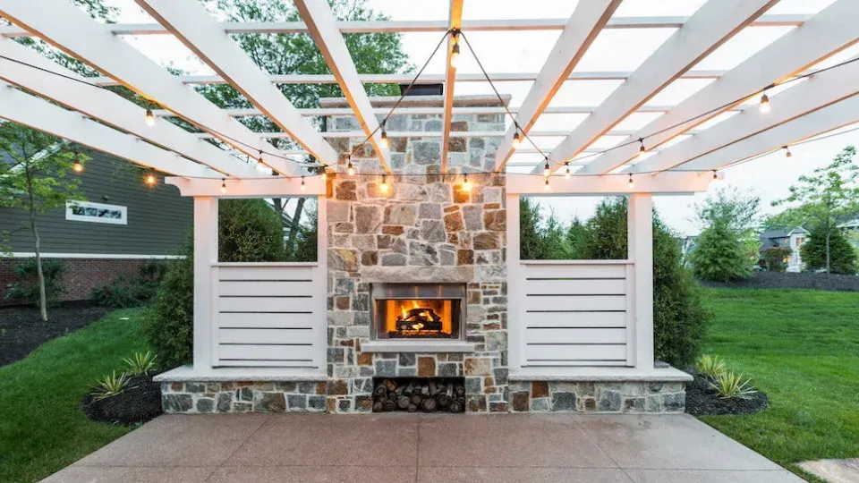 Outdoor fireplace with stone facade, under a white pergola with string lights, patio, green grass. Outdoor fireplace with stone facade, under a white pergola with string lights, patio, green grass.