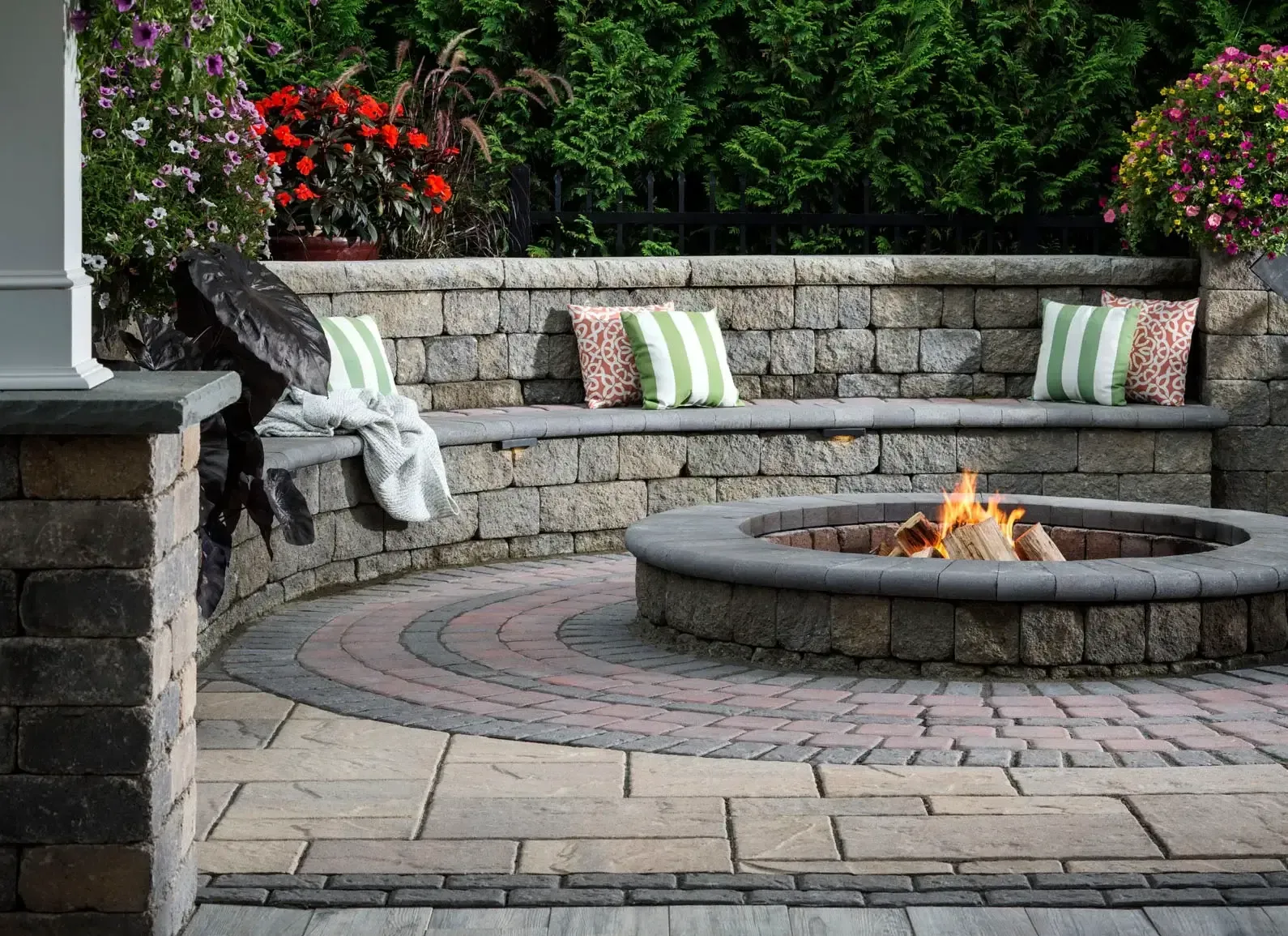 Stone patio with built-in bench and fire pit, green and pink patterned pillows, flowers, and a blank