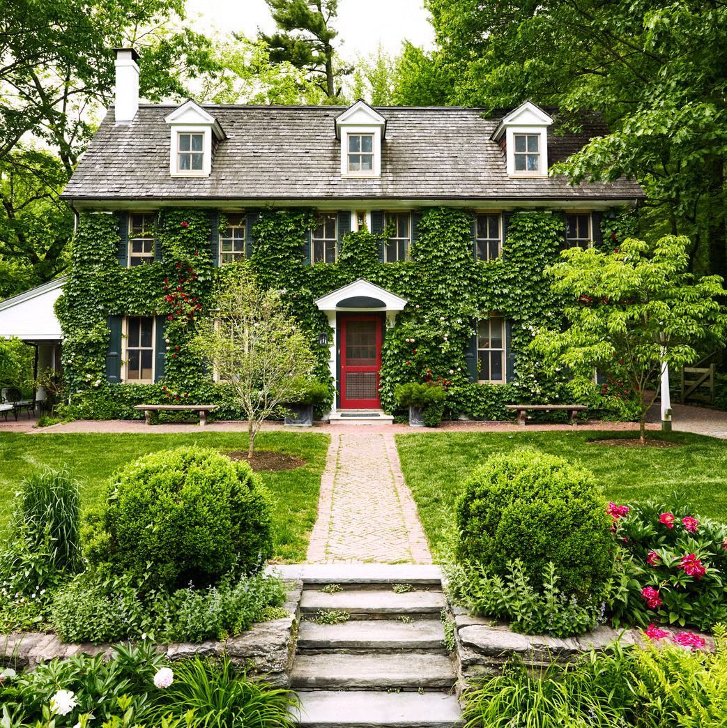 Two-story house with red door covered in ivy. Stone walkway, green lawn, garden with bushes and flowers.