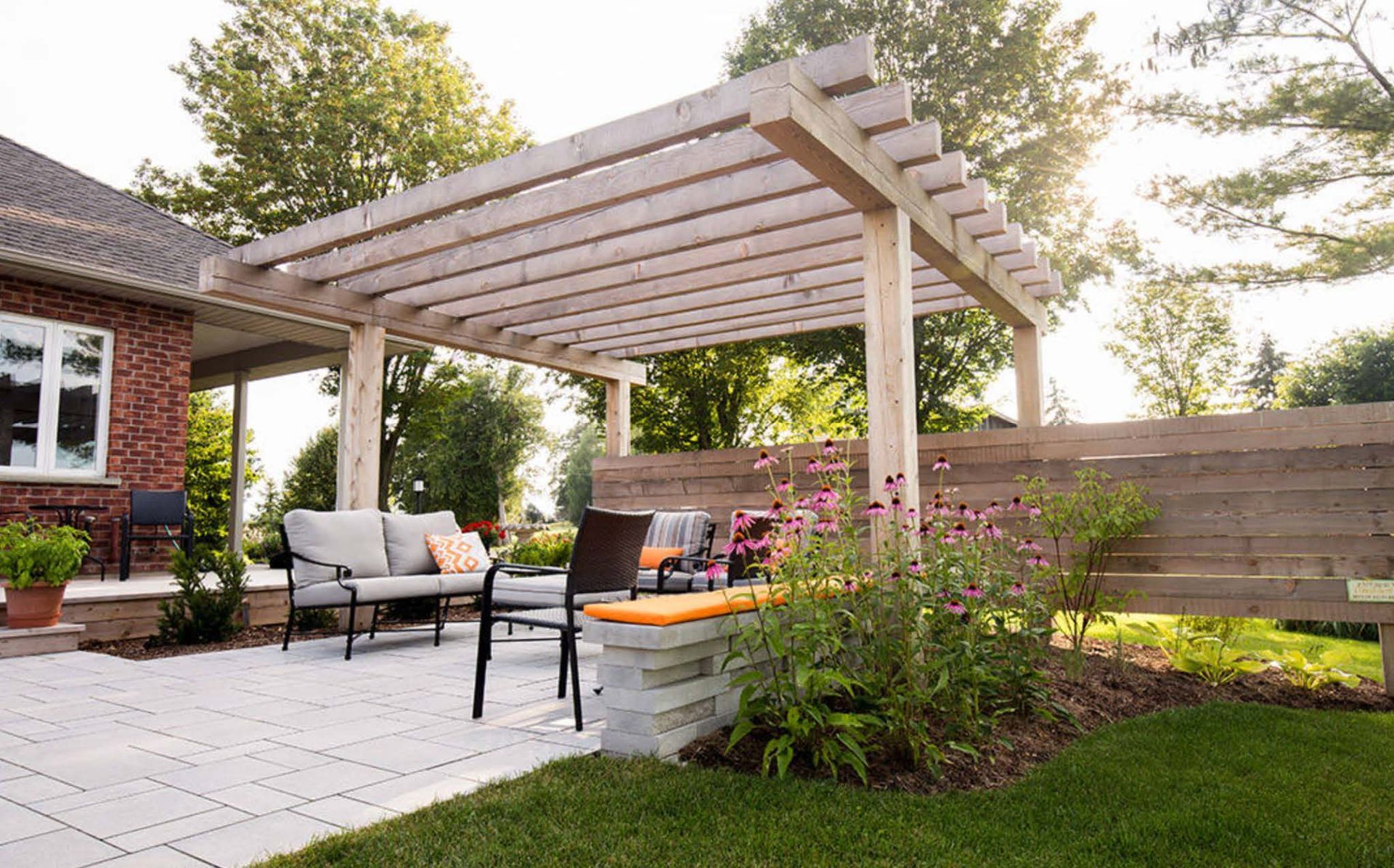 Patio with pergola, outdoor seating, and landscaping next to a red brick house.