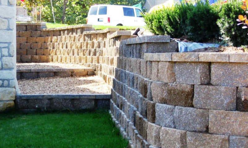 Stone retaining walls with steps leading up, gravel, and grass. A van and bushes are in the background.