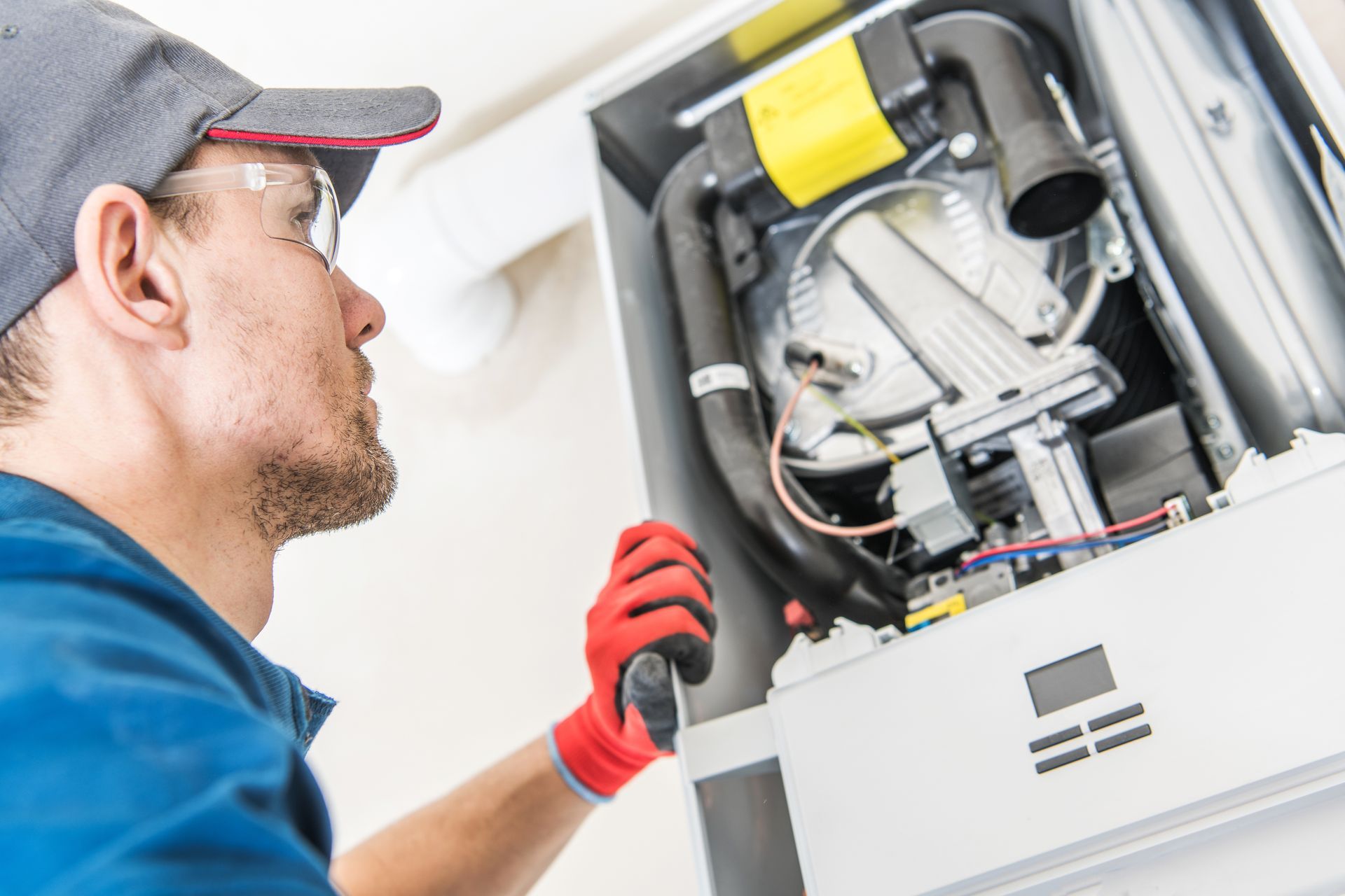 A technician in a hat and gloves repairs a white furnace.