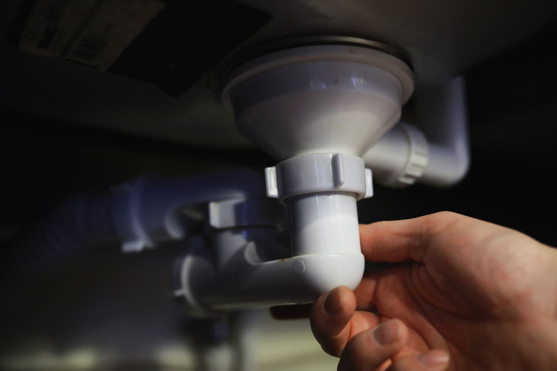 Hand adjusting a white plastic sink drain pipe under a dark sink.