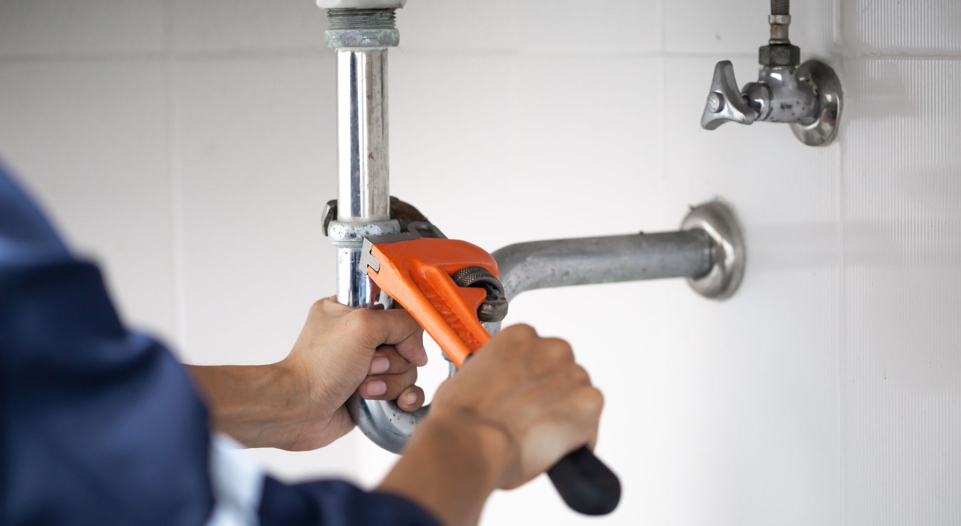 Plumber using an orange wrench to tighten a pipe under a sink in a white-tiled bathroom.