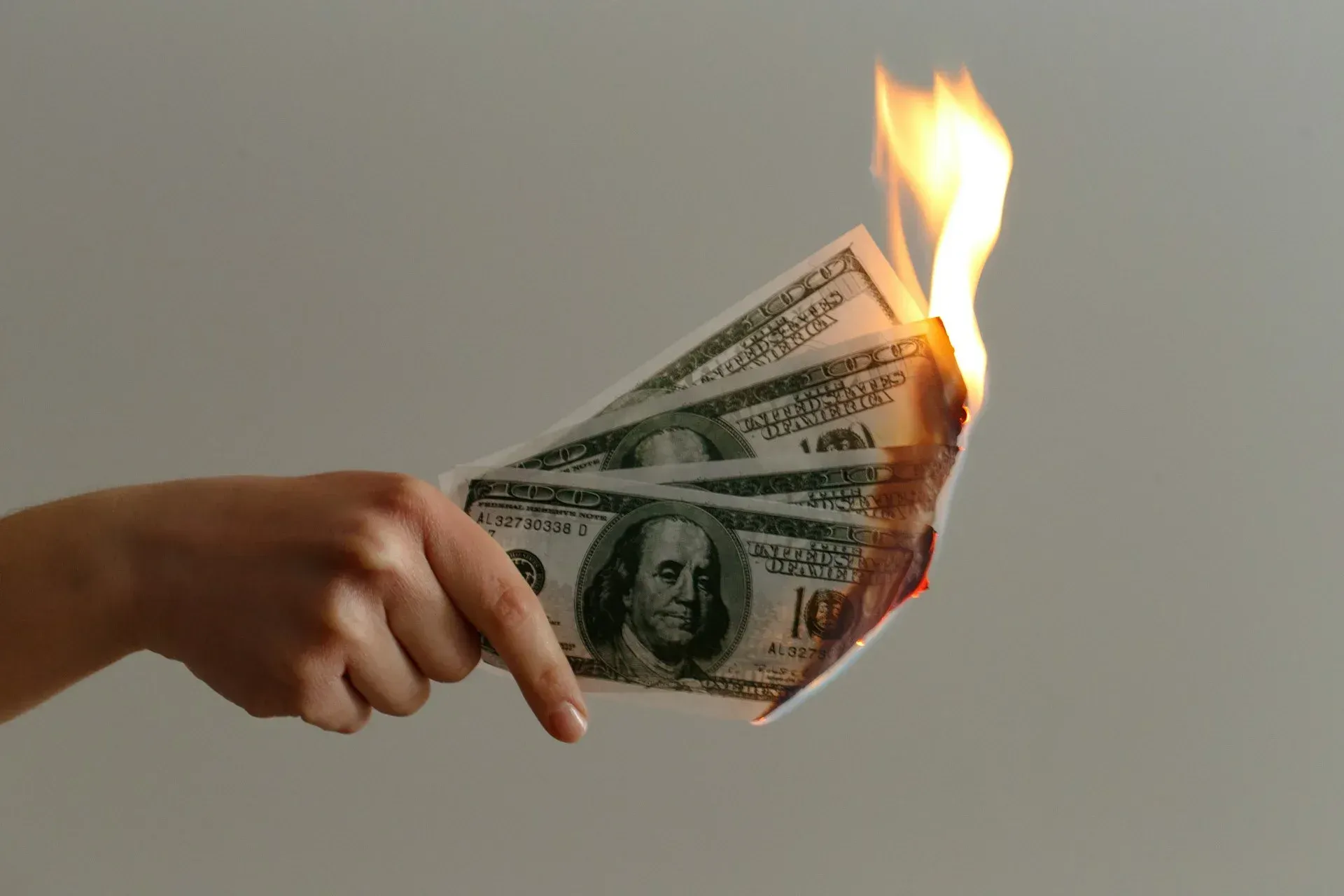 Hand holding burning US $100 bills, flames and smoke visible.